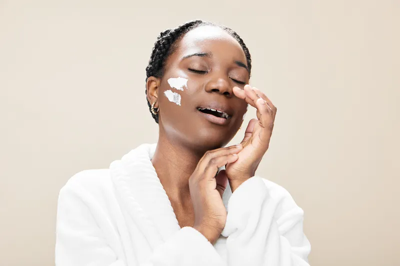 Beautiful young Black woman applying skincare cream, demonstrating a healthy skincare routine, with joy and relaxation against a light beige background
