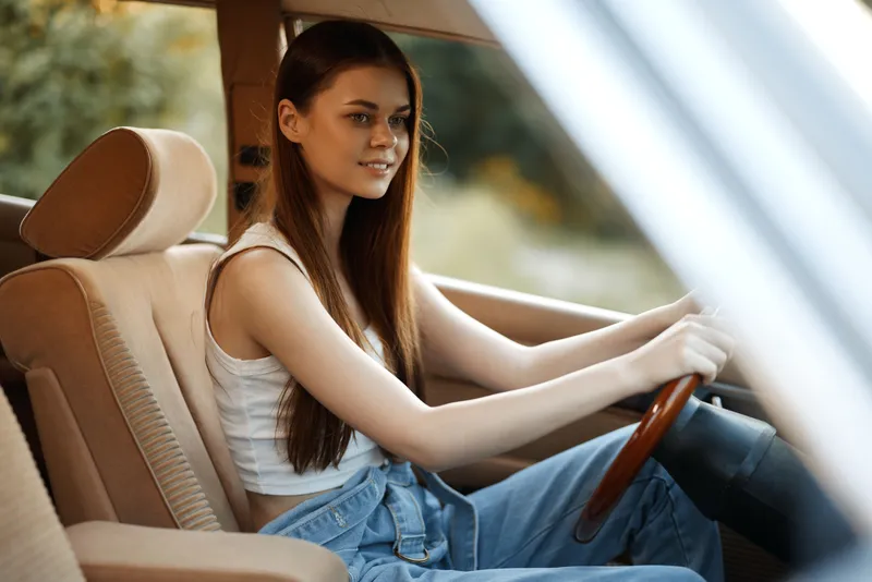 Young woman driving vintage car with long hair, wearing casual outfit, showcasing confidence and joy against natural background.