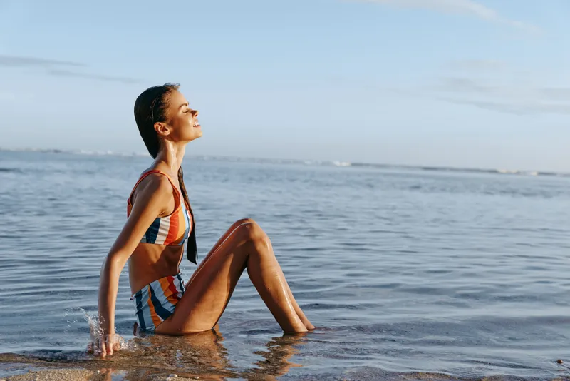 Woman sitting in shallow ocean water on a beach, wearing a striped swimsuit, calm summer relaxation, profile view, soft natural light, sea horizon in background, outdoor lifestyle