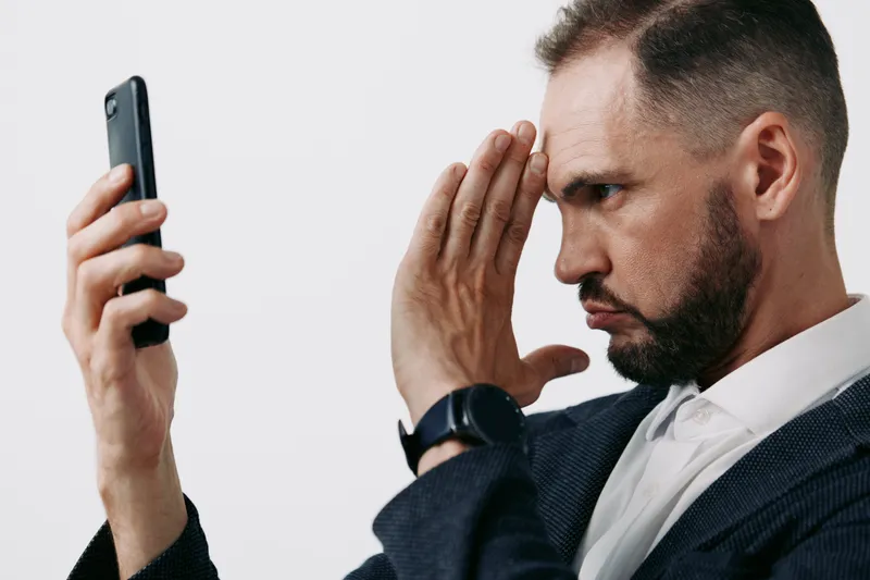 A focused business professional man poses with a smartphone, signaling contemplation against an isolated color background, capturing subtle emotions of concentration and concern.