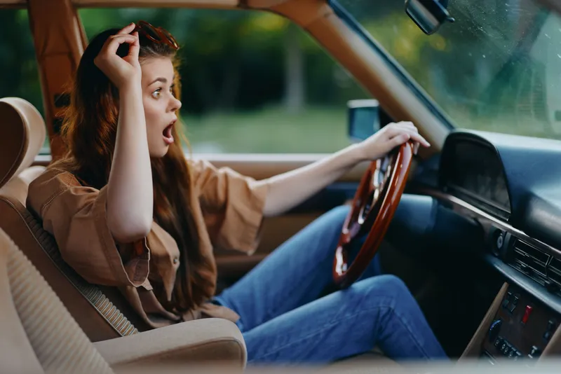 Surprised young woman driving in a vintage car, expressing excitement and adventure, surrounded by greenery in a vibrant, sunny setting.