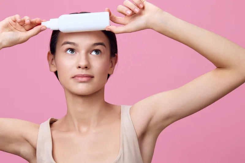 Young woman with short hair holding a skincare bottle over her forehead against a pastel pink background, radiating confidence and beauty