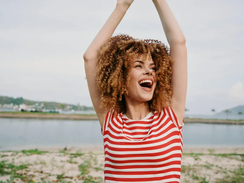Joyful woman with curly hair wearing a red and white striped shirt raises her arms in laughter beside a calm lake, radiating carefree summer energy.