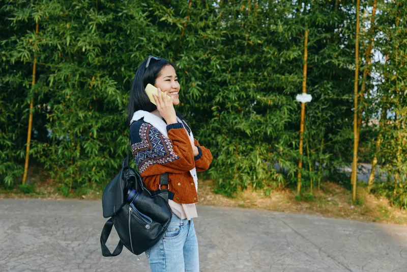 Woman with backpack talking on smartphone in an outdoor park setting, casual fashion and accessories, trees and pavement in the background, candid urban lifestyle moment