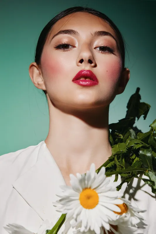 Close-up portrait of a young woman with glossy red lips and soft pink blush holding white daisies against a muted teal background. Vibrant beauty and creative fashion concepts.