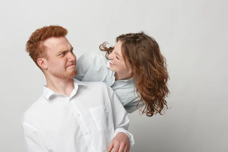 Redhead man in white shirt looks surprised while woman with curly hair playfully leans on his shoulder. Studio portrait on grey background with casual clothing and friendly interaction concept.