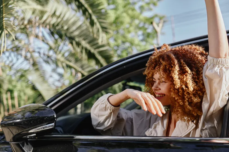 smiling woman with curly hair enjoying a sunny day inside a black car, vibrant tropical background with green palm trees and relaxed summer vibe