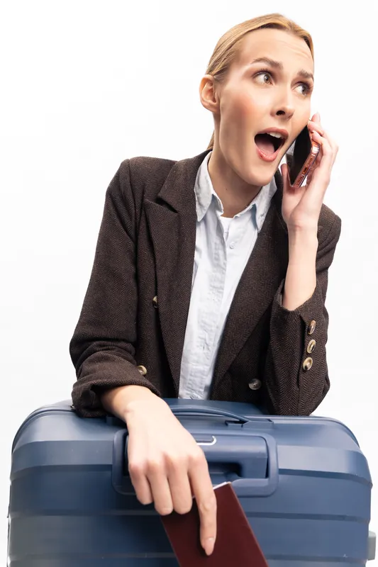 Surprised businesswoman with suitcase holds passport and talks on phone. Studio portrait on white background. Concept of travel, business trip, communication, and urgent news.