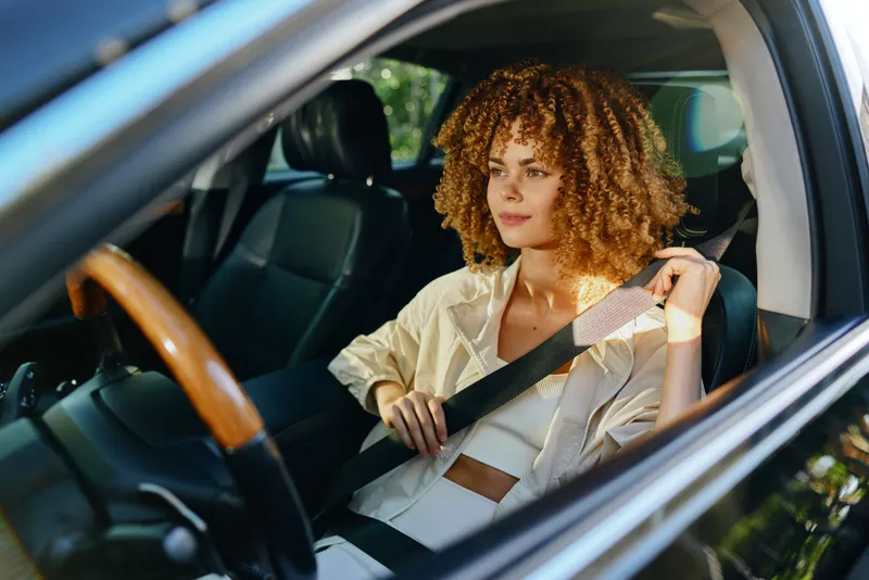 Young woman with curly hair fastening seatbelt inside a modern car, wearing casual light clothing, demonstrating safety awareness and preparedness.