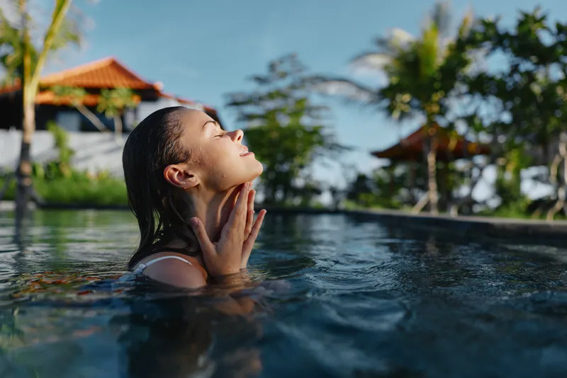 Young woman with wet hair relaxes in a sunlit pool, her face tilted toward the light as water ripples softly around her, a moment of tranquil summer bliss.