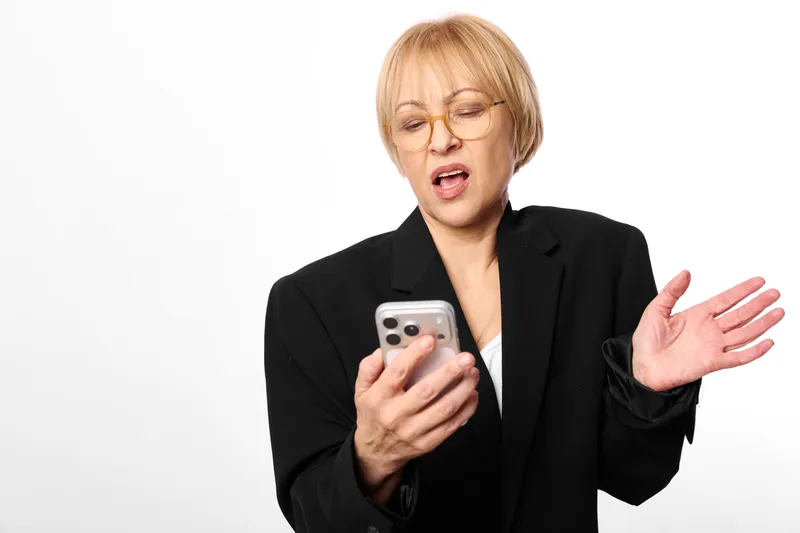Stressed woman with smartphone and glasses, hand raised as she looks shocked and frustrated, isolated on white background, concept for anxiety, panic, communication overload today