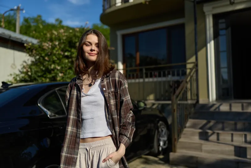 Casual woman outdoors on a sunny day, wearing a plaid shirt over a white top, standing in front of a modern house, embodying a relaxed lifestyle moment