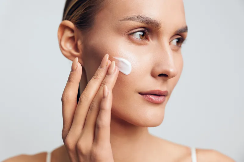 Young woman with clear skin applies moisturizer cream on her cheek while looking thoughtfully to the side against a plain light background.