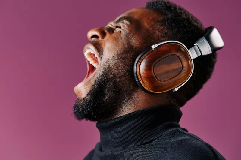 African man expressing joy while singing loudly with wooden headphones against a vibrant purple background, showcasing emotions and a modern lifestyle