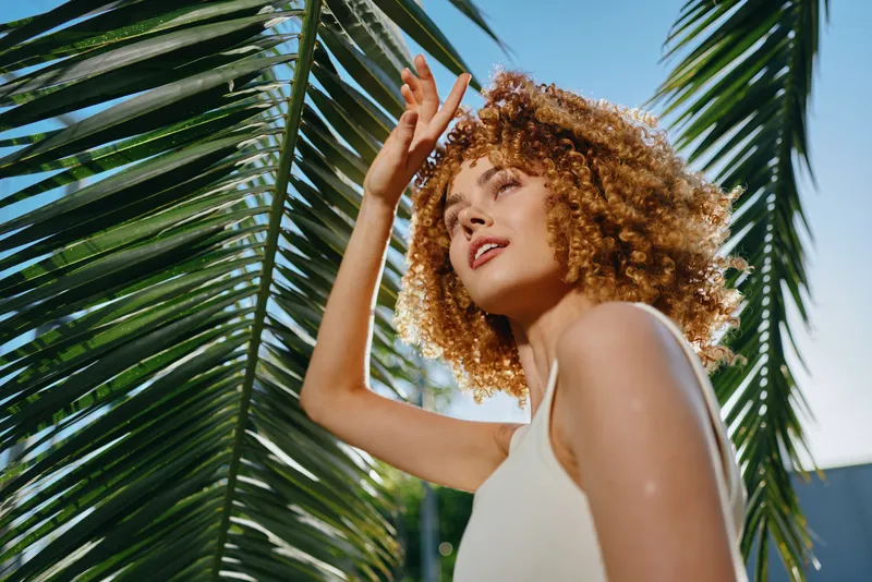 smiling woman with curly hair enjoys sunny tropical day near large palm leaves wearing white tank top in bright outdoor setting