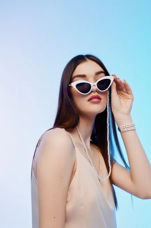 Young woman in stylish white sunglasses and beige dress poses confidently against a light blue background. Concept of woman beauty fashion and elegance.