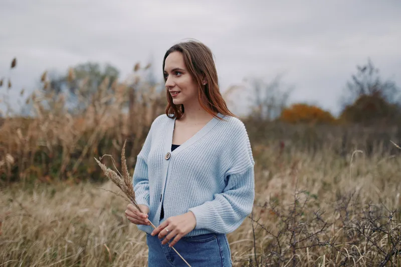 A candid portrait of a thoughtful woman in a soft blue knit cardigan standing in a sunlit field, conveying authenticity and natural charm.