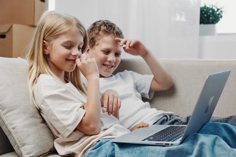 Two happy children sitting on a couch using a laptop together in a bright living room. Concept of childhood friendship, technology use, and home comfort.