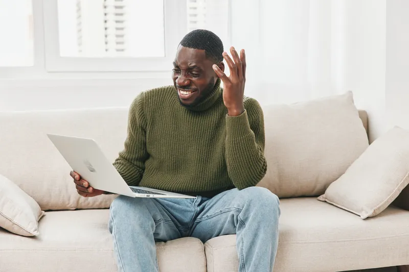 Happy black man using laptop at home, expressing joy while working or exploring online content, wearing a green sweater, casual home environment