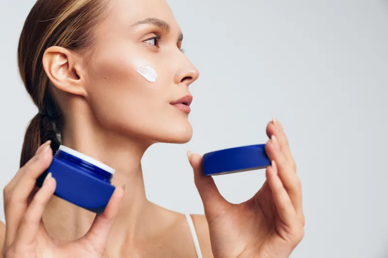 Bright close-up of a woman applying moisturizer, cream on her cheek, soft lighting, skincare routine in progress, natural makeup, calm expression, fresh glow, blue jars