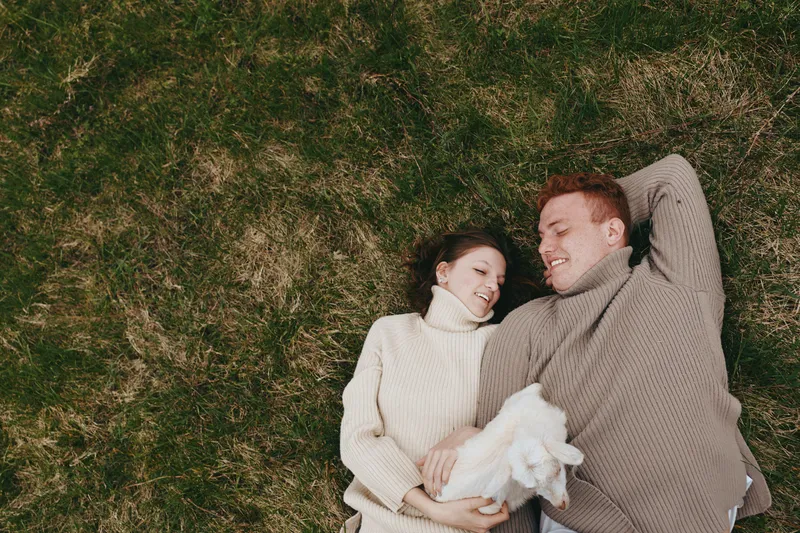 A couple lying on grass wearing knit sweaters, cradling a baby in a soft blanket, sharing a tender moment outdoors. Lifestyle scene focused on family warmth, bonding, and gentle affection under