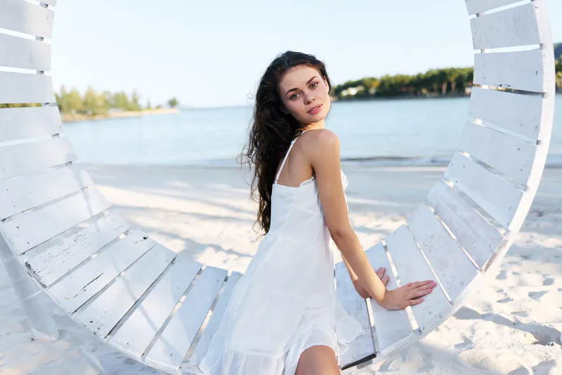 A young woman in a flowing white dress stands on a sunlit beach, framed by a circular white structure. The sea, sand and sky create a calm, airy mood.