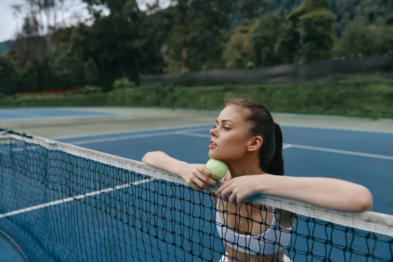 Young athletic woman in sportswear leaning on tennis net, holding a tennis ball, demonstrating focus and determination on a sunny day in a green outdoor setting