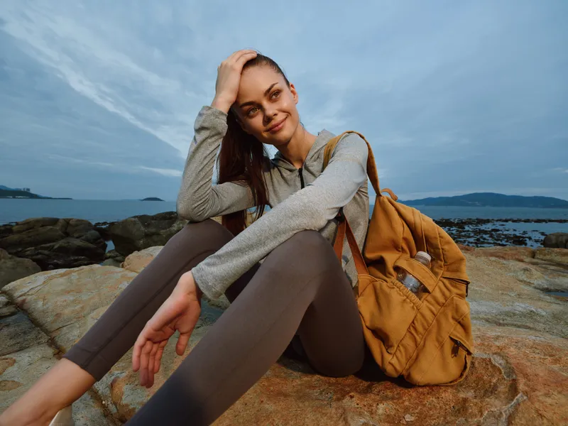 Young woman enjoying travel on rocky beach at sunset, wearing casual sports outfit, smiling with backpack, serene atmosphere, outdoor adventure, nature vibes