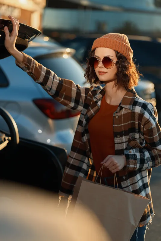Woman shopping at car trunk with bag, beanie and sunglasses in plaid jacket, candid lifestyle portrait capturing authenticity and golden hour glow, mindful living and emotional storytelling.