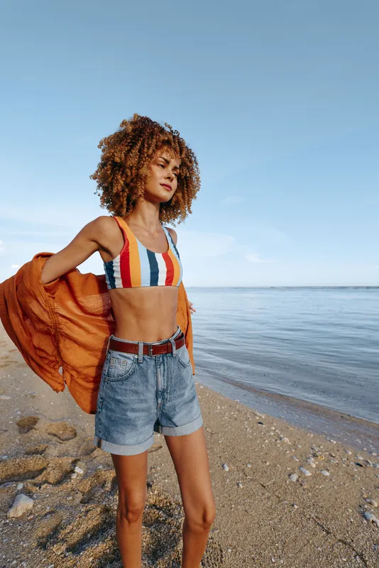Young woman with curly hair wears striped top and denim shorts on a beach by the sea.