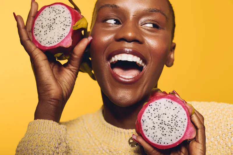 Happy African woman with dark skin holds two halves of dragon fruit near face, smiling widely on yellow background. Concept of healthy lifestyle, natural food, and joyful emotions.