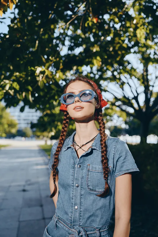 Lifestyle woman in modern boho-western denim street style wearing blue sunglasses and red hair accessories in film color outdoor setting under green trees.