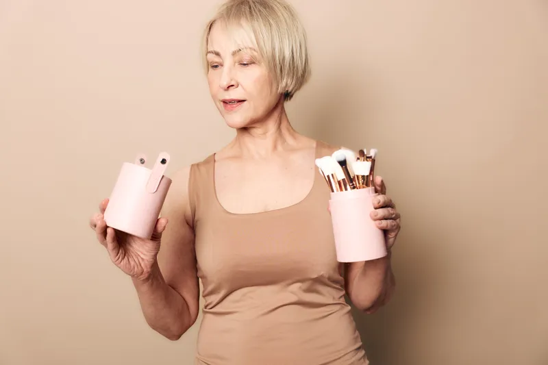 Mature woman with short blonde hair holding pink containers with makeup brushes on beige background. Concept of beauty tools, cosmetics, skincare routine, and personal grooming at home.