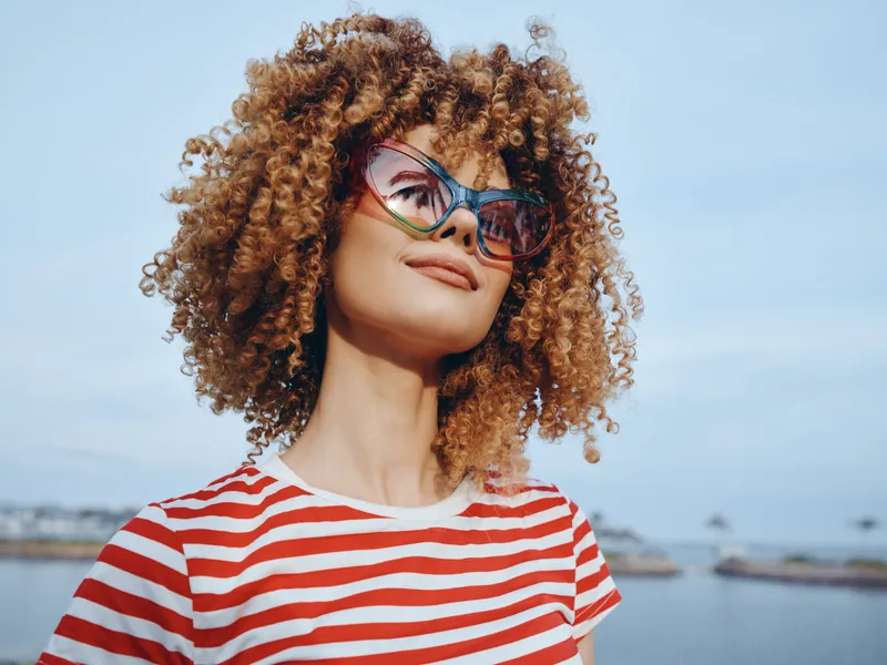 Portrait of a smiling woman with curly hair, wearing a red and white striped shirt and oversized sunglasses against a sunny seaside backdrop, radiating confidence and carefree style.