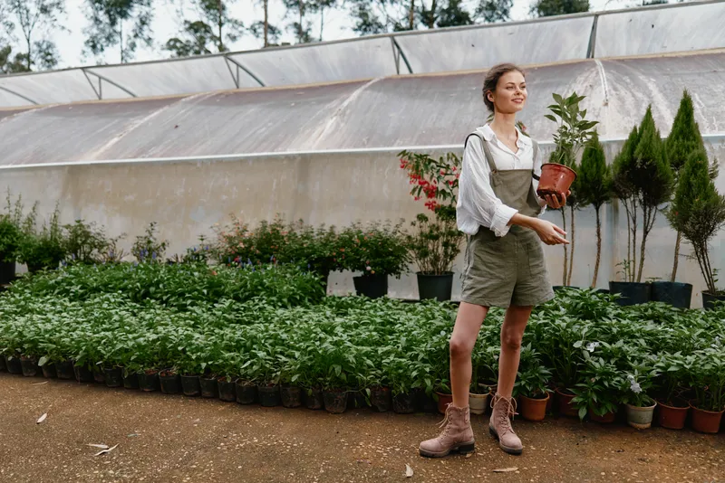 Young woman holding a potted plant in a greenhouse, wearing casual overalls and boots, surrounded by lush greenery, embodying a love for nature and sustainable living