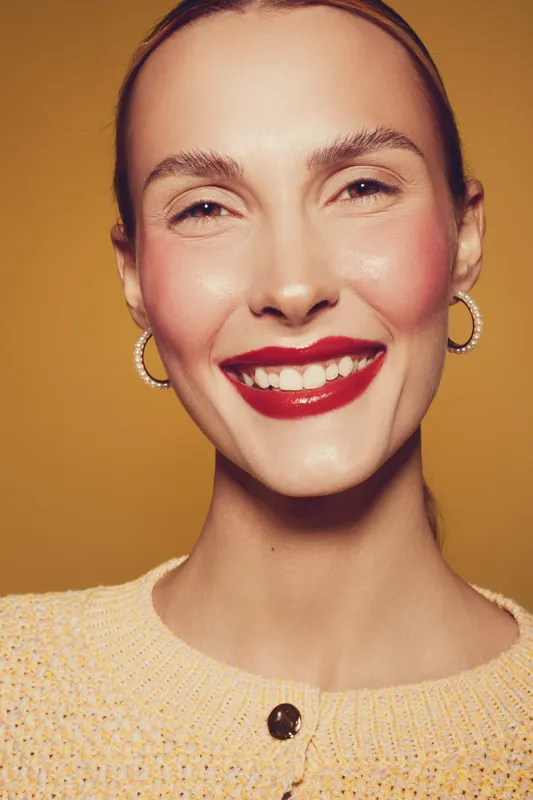 Smiling woman with red lipstick and hoop earrings poses against yellow background. Close-up portrait shows natural skin texture and joyful expression. Concept of beauty and happiness.