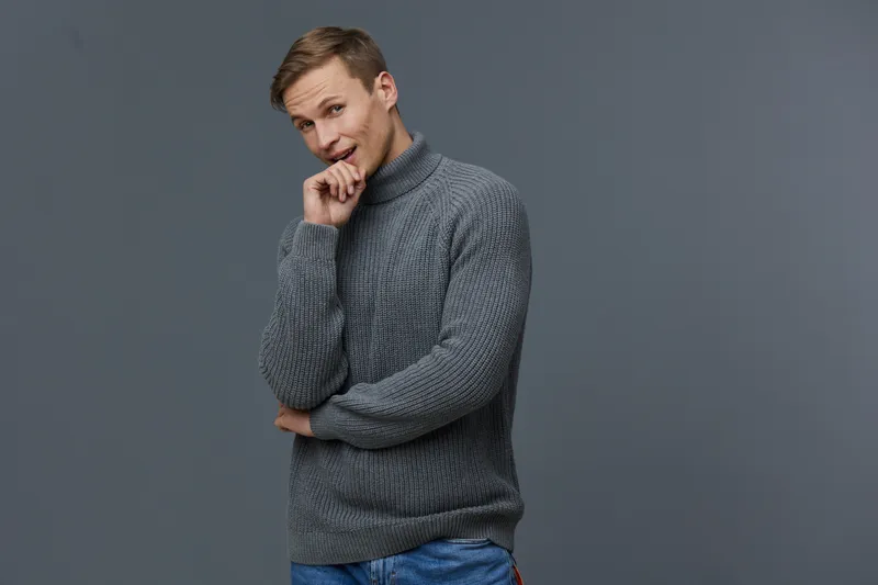 Casual young man wearing gray sweater and blue jeans posing against plain gray background. Smiling, thoughtful, confident young male model with friendly expression, relaxed stance, modern fashion look