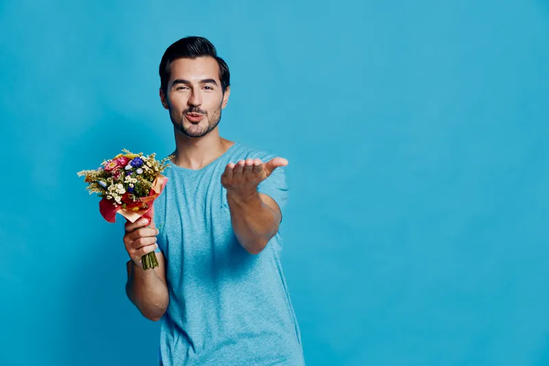Charming smiling man holding a bouquet of colorful flowers, blowing a kiss towards the camera. Bright blue background enhances the cheerful mood.
