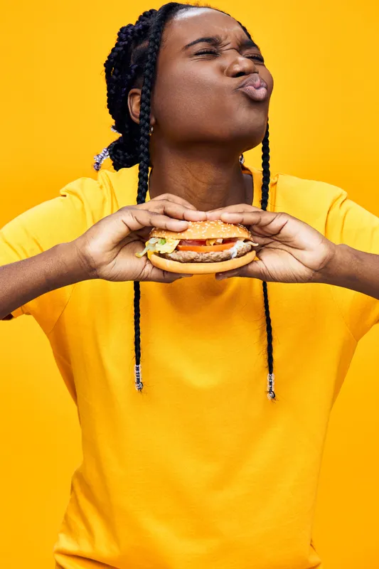 Happy young Black woman enjoying a colorful burger against a vibrant yellow background, showcasing joy and a love for fast food