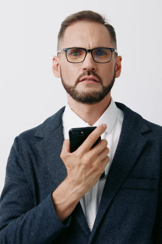 A professional man stands against a colored isolated background, expressing focus and contemplation while holding a smartphone, portraying business confidence and clear intent.