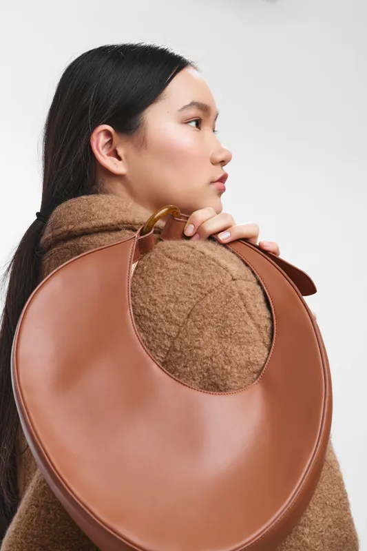 Asian woman wearing a warm brown coat shows a side profile as she carries a round leather bag over her shoulder, creating a chic and calm fashion moment.