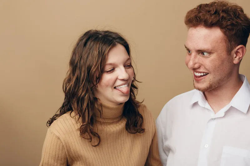 Young woman in brown sweater playfully sticks out tongue at smiling redhead man in white shirt. Friendly interaction and casual mood on beige background. Concept of friendship and fun.