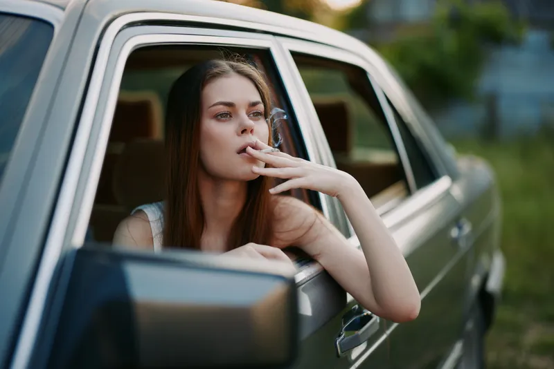 A young Caucasian woman smoking while relaxing in a car, showcasing a calm expression and casual summer attire, surrounded by a lush green background.