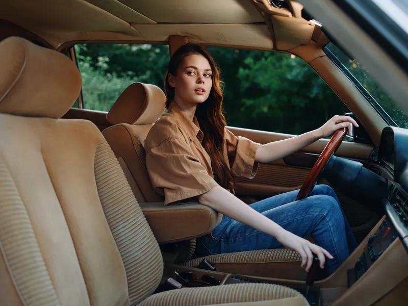 Young woman in a stylish brown shirt driving a vintage car, exuding confidence and freedom, set against a lush green background.