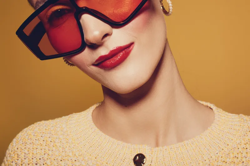 Close-up of a stylish woman wearing oversized red sunglasses and red lipstick. She wears a yellow knitted sweater and gold hoop earrings on a warm yellow background. Fashion concept.