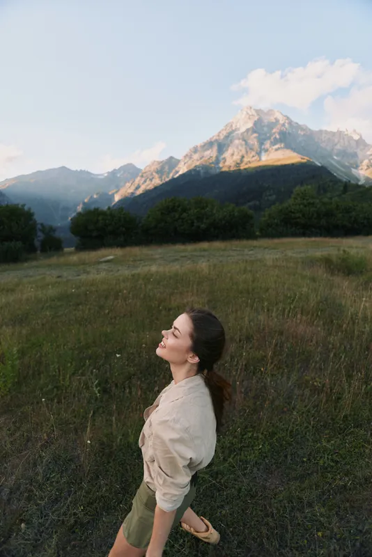 Young woman wearing a beige blouse and shorts stands in a grassy meadow, gazing toward distant mountains at sunset, with a calm, hopeful mood and a sense of freedom.