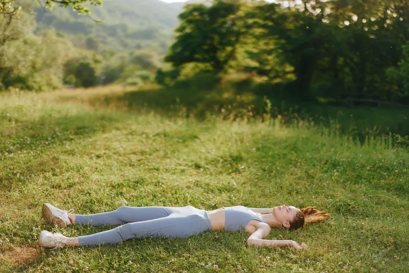 Young woman in activewear relaxing on the grass in a serene natural setting, capturing the essence of tranquility and mindfulness in outdoor fitness activities