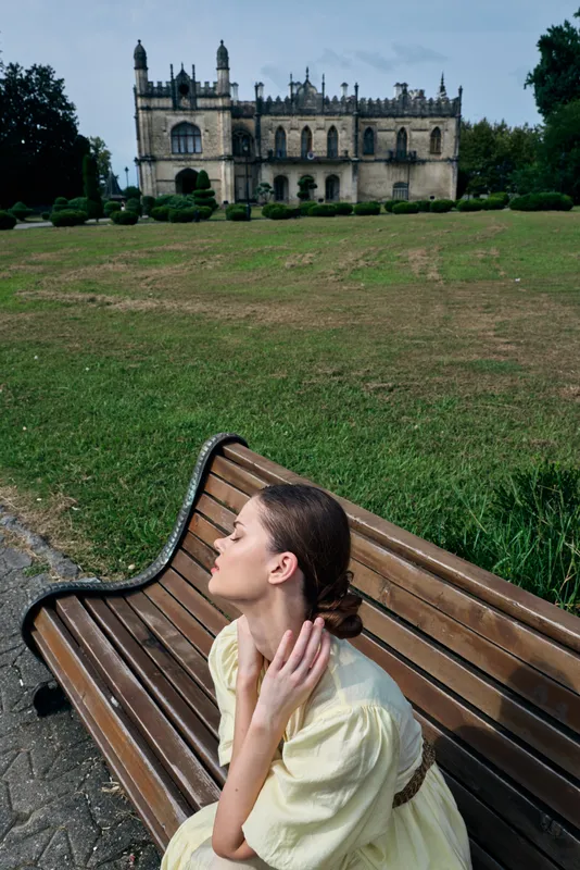 woman sits on a wooden bench in a lush park, gazing upward as sunlight highlights a distant castle, creating a serene moment of outdoor scenery and quiet reflection