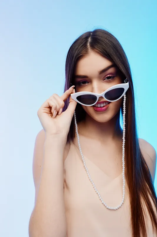 Young woman with long hair wearing white sunglasses and a beige top poses against a blue background. Concept of woman beauty fashion and modern style.