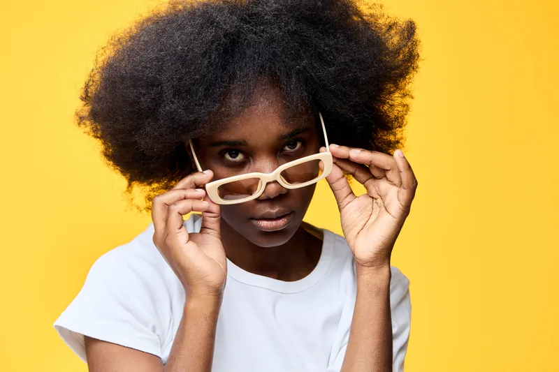 Young African woman with curly hair wearing stylish glasses, looking confidently at the camera against a bright yellow background